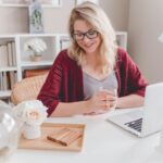 Woman smiling holding glass mug sitting beside table with macbook